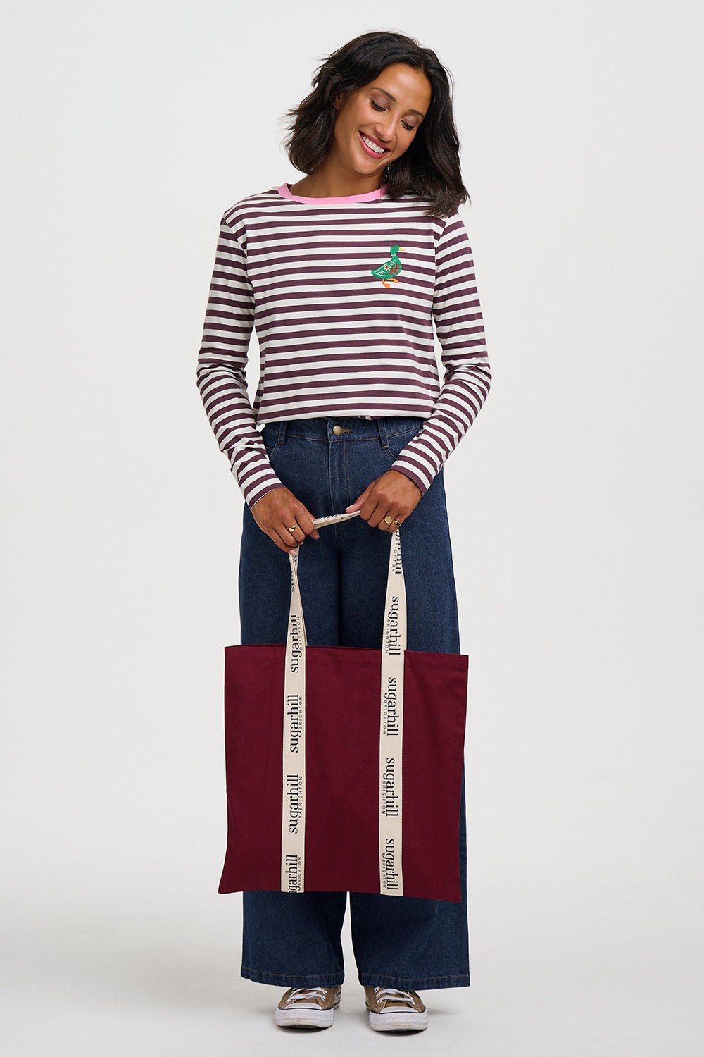 A woman modeling a long-sleeve striped shirt with a small green embroidered accent, paired with wide-leg blue jeans and holding a burgundy tote bag with white handles against a light gray background.
