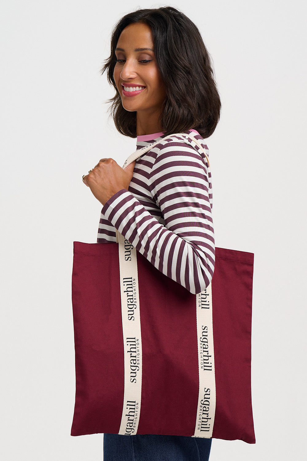 A woman wearing a burgundy and white striped long-sleeve shirt holds a large burgundy tote bag with cream-colored shoulder straps printed with the brand name 'sugarhill' in black.