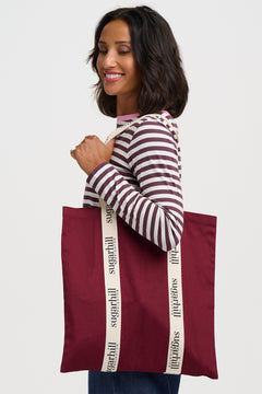 A woman wearing a burgundy and white striped long-sleeve shirt holds a large burgundy tote bag with cream-colored shoulder straps printed with the brand name 'sugarhill' in black.