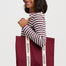 A woman wearing a burgundy and white striped long-sleeve shirt holds a large burgundy tote bag with cream-colored shoulder straps printed with the brand name 'sugarhill' in black.