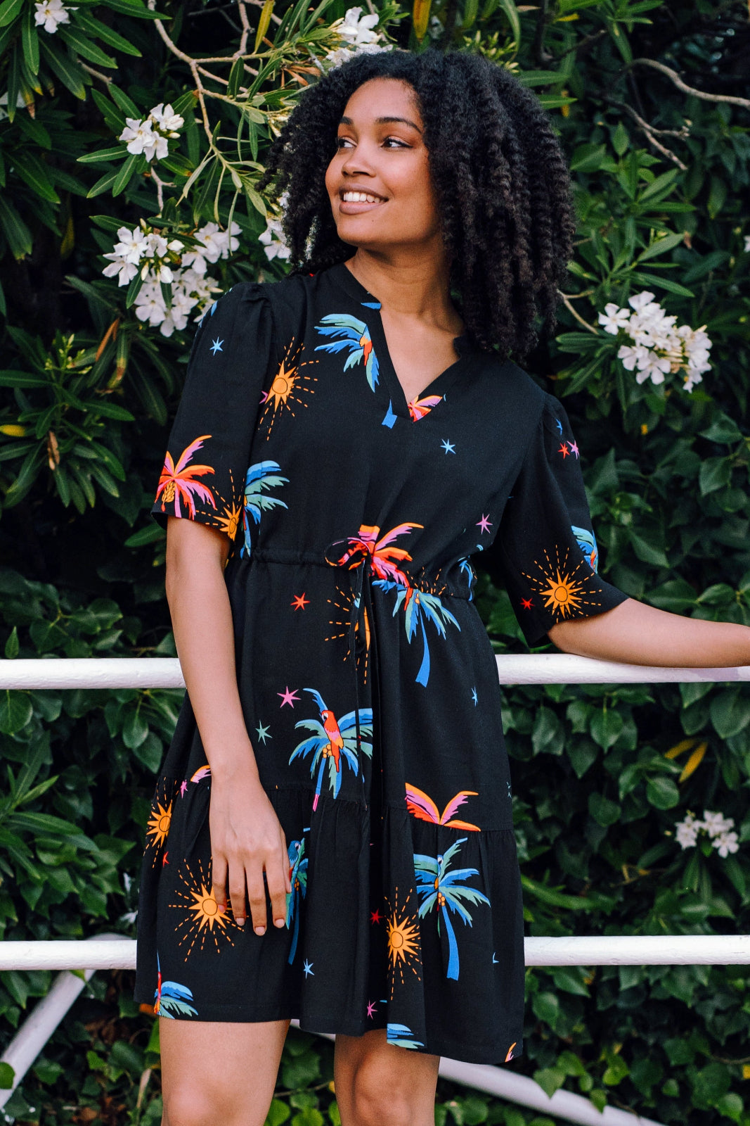 A woman wearing a short-sleeved, tiered mini black dress with a palm tree and sunset print, standing near greenery.