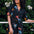 A woman wearing a short-sleeved, tiered mini black dress with a palm tree and sunset print, standing near greenery.