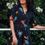 A woman wearing a short-sleeved, tiered mini black dress with a palm tree and sunset print, standing near greenery.