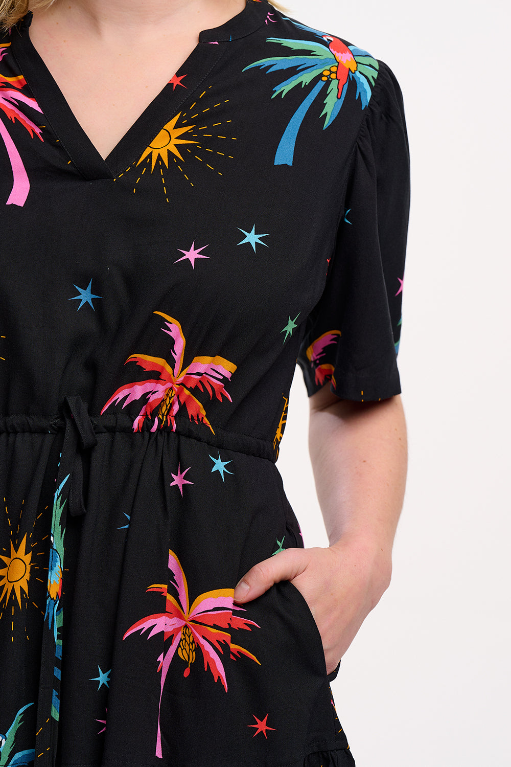 A model wearing a short-sleeved mini dress with a deep V-neck cut-out, featuring a tiered skirt, palm tree, and other colorful patterns on a black background.