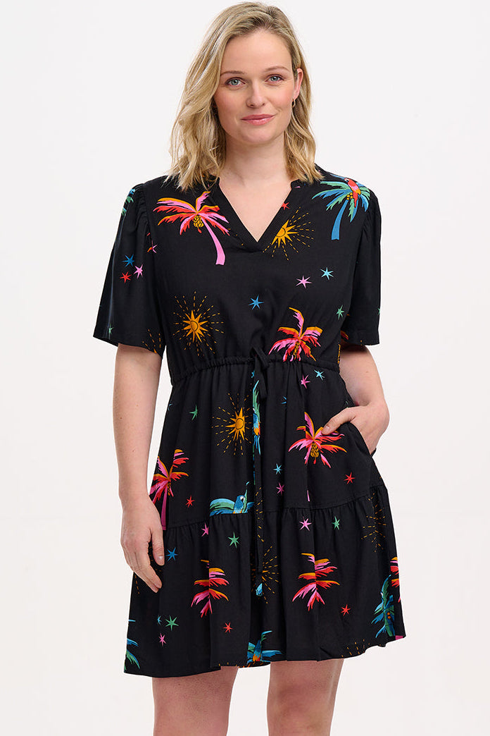 A model wearing a short-sleeved mini dress with a deep V-neck cut-out, featuring a tiered skirt, palm tree, and other colorful patterns on a black background.
