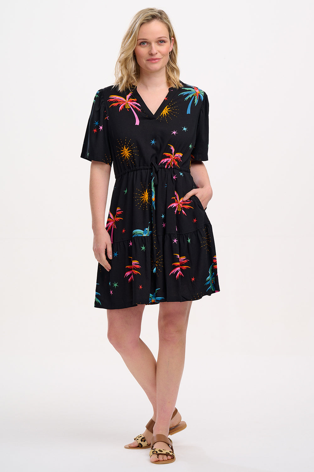A model wearing a short-sleeved mini dress with a deep V-neck cut-out, featuring a tiered skirt, palm tree, and other colorful patterns on a black background.