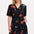 A model wearing a short-sleeved mini dress with a deep V-neck cut-out, featuring a tiered skirt, palm tree, and other colorful patterns on a black background.