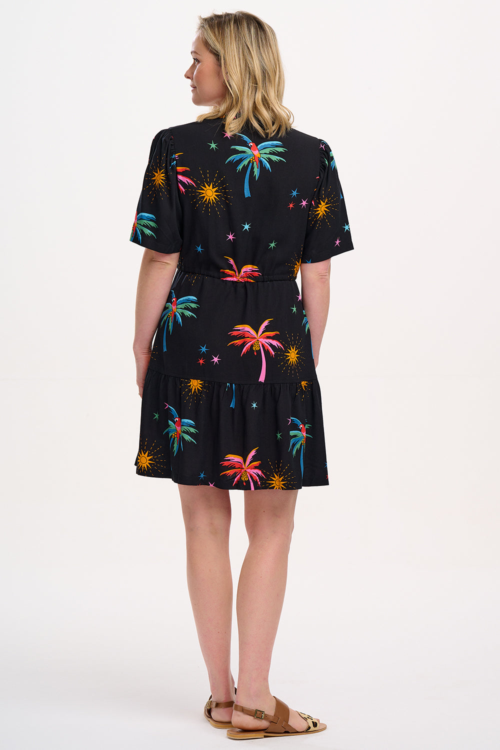 A model wearing a short-sleeved mini dress with a deep V-neck cut-out, featuring a tiered skirt, palm tree, and other colorful patterns on a black background.