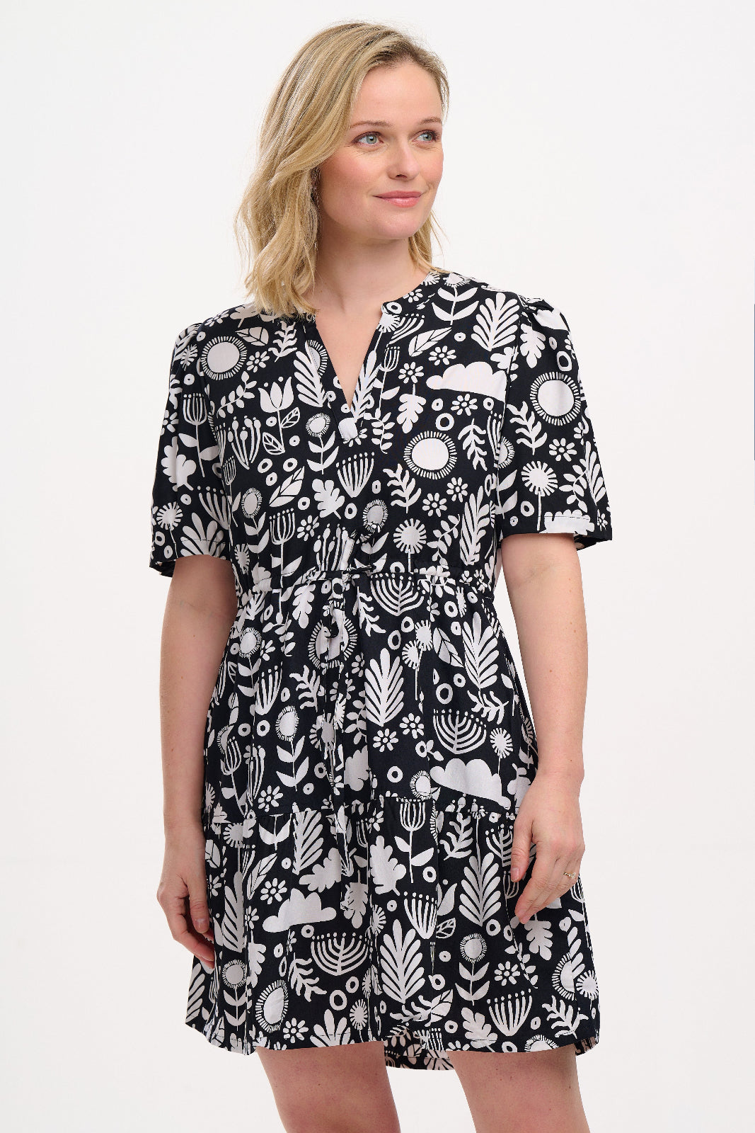 A woman wearing a knee-length V-neck dress with short sleeves and a tiered skirt. The dress features a black and white floral folk print.