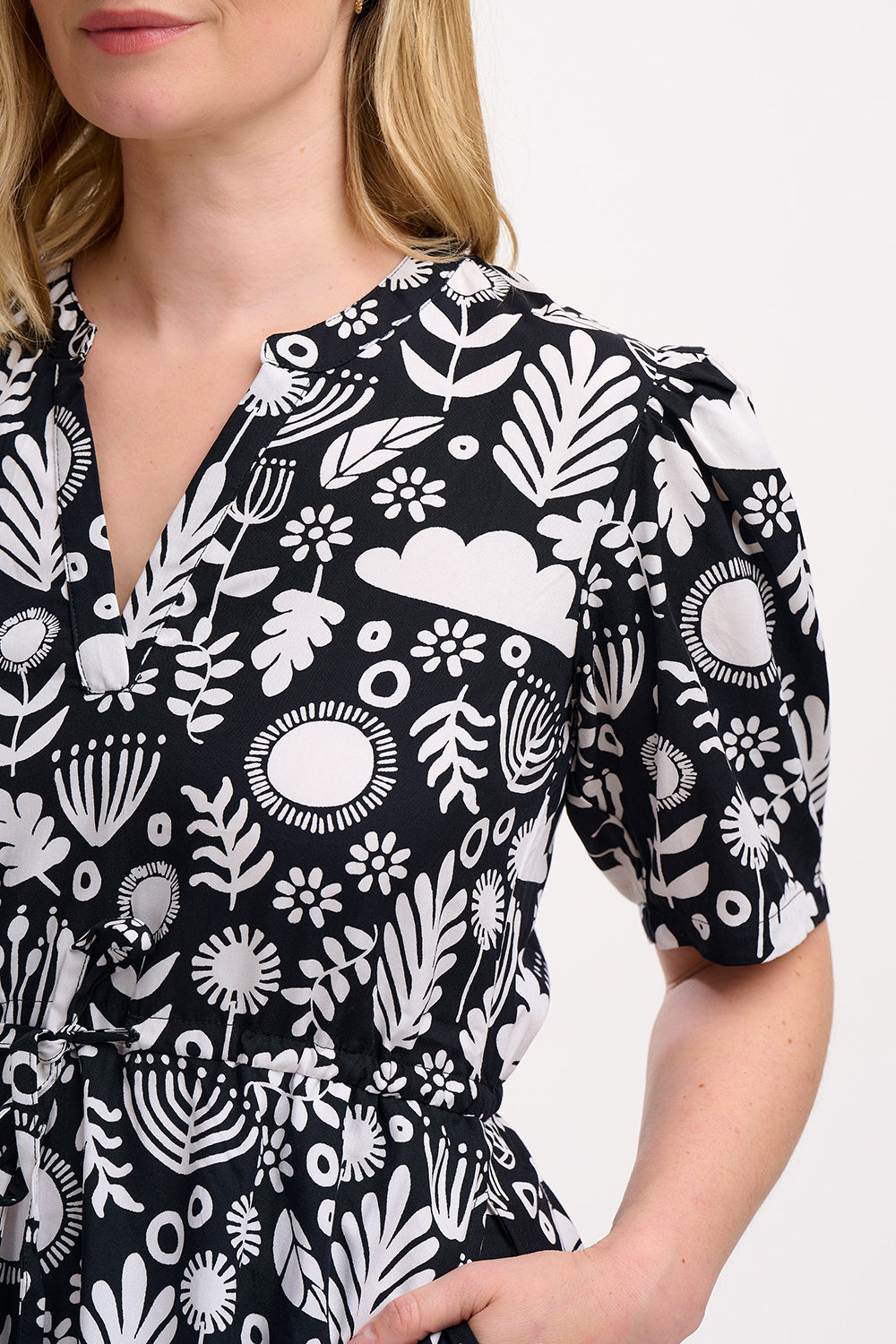 A woman wearing a knee-length V-neck dress with short sleeves and a tiered skirt. The dress features a black and white floral folk print.
