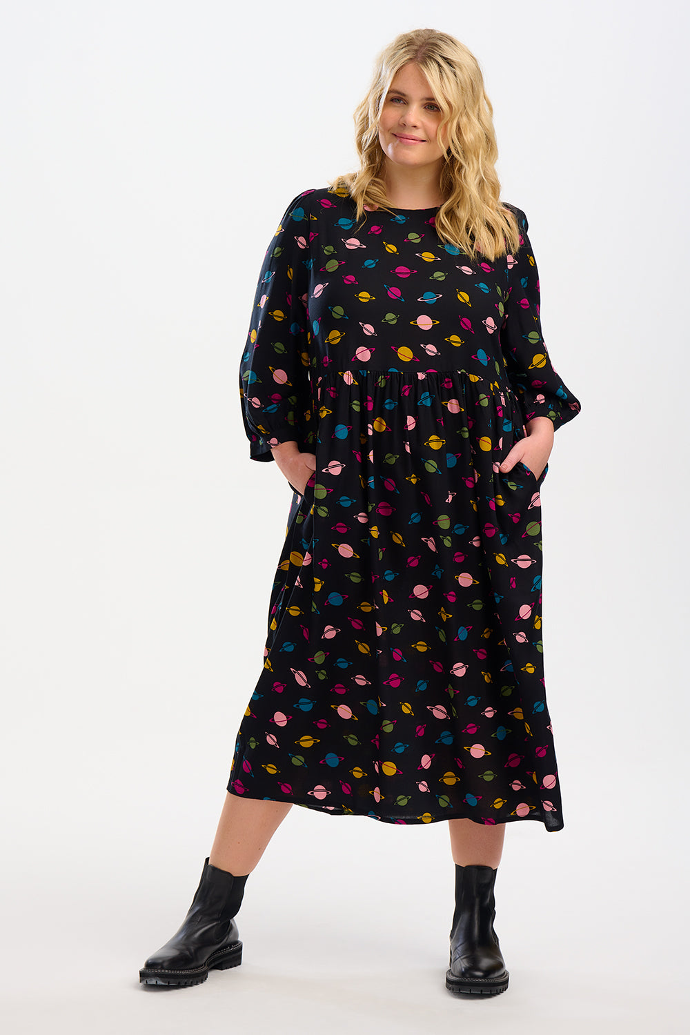 A woman standing, wearing a midi smock dress with a colorful planet pattern on a black background, paired with black boots.