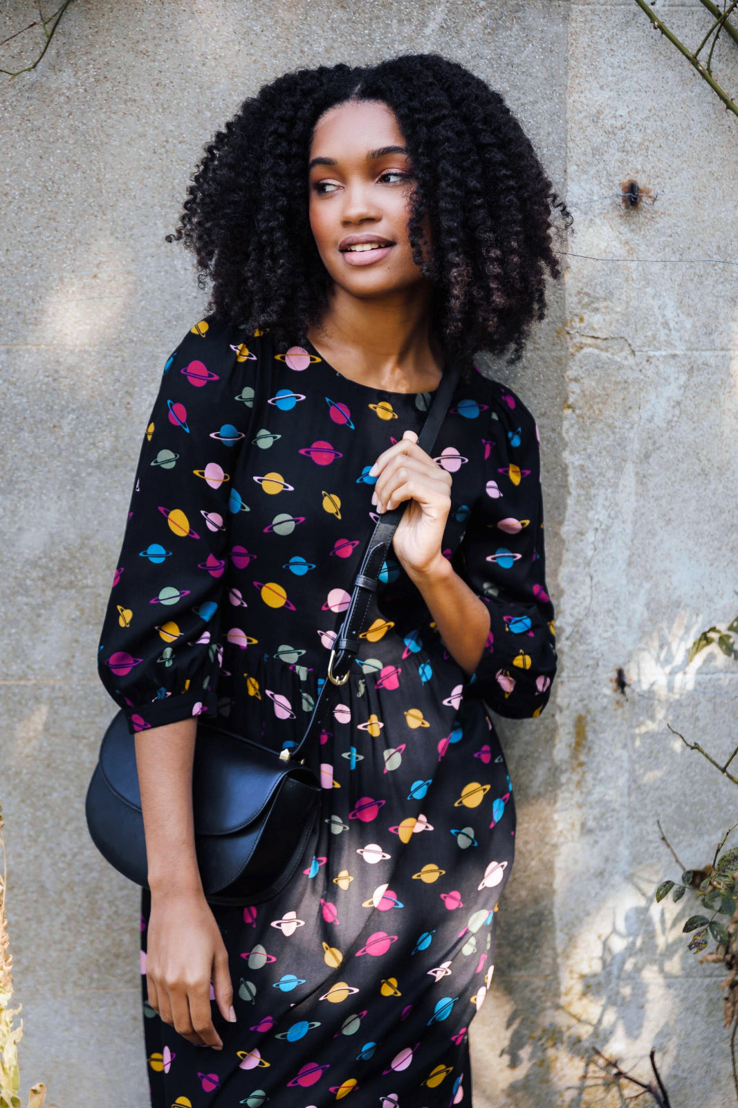 A woman standing, wearing a midi smock dress with a colorful planet pattern on a black background, paired with black boots.
