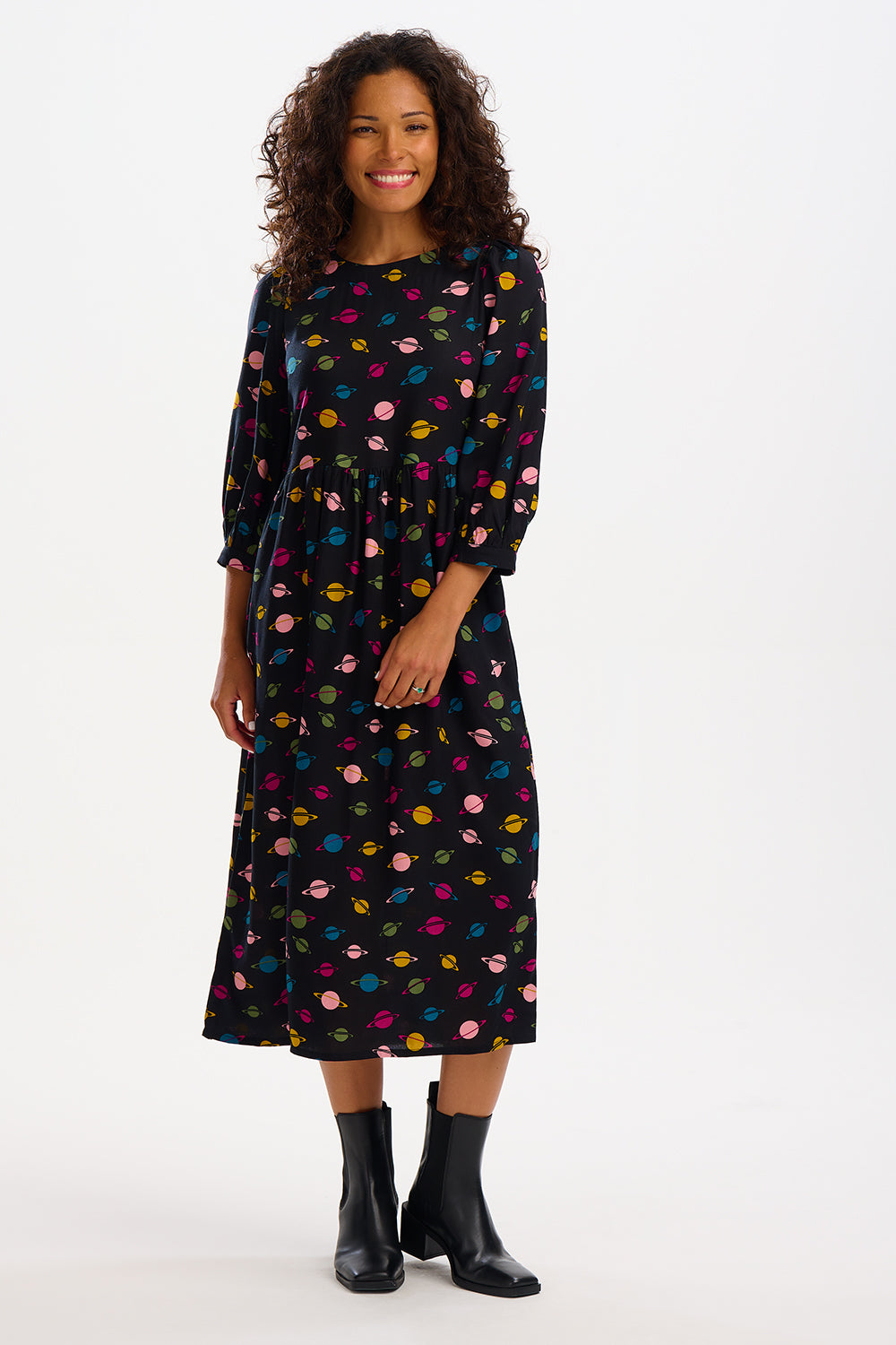 A woman standing, wearing a midi smock dress with a colorful planet pattern on a black background, paired with black boots.
