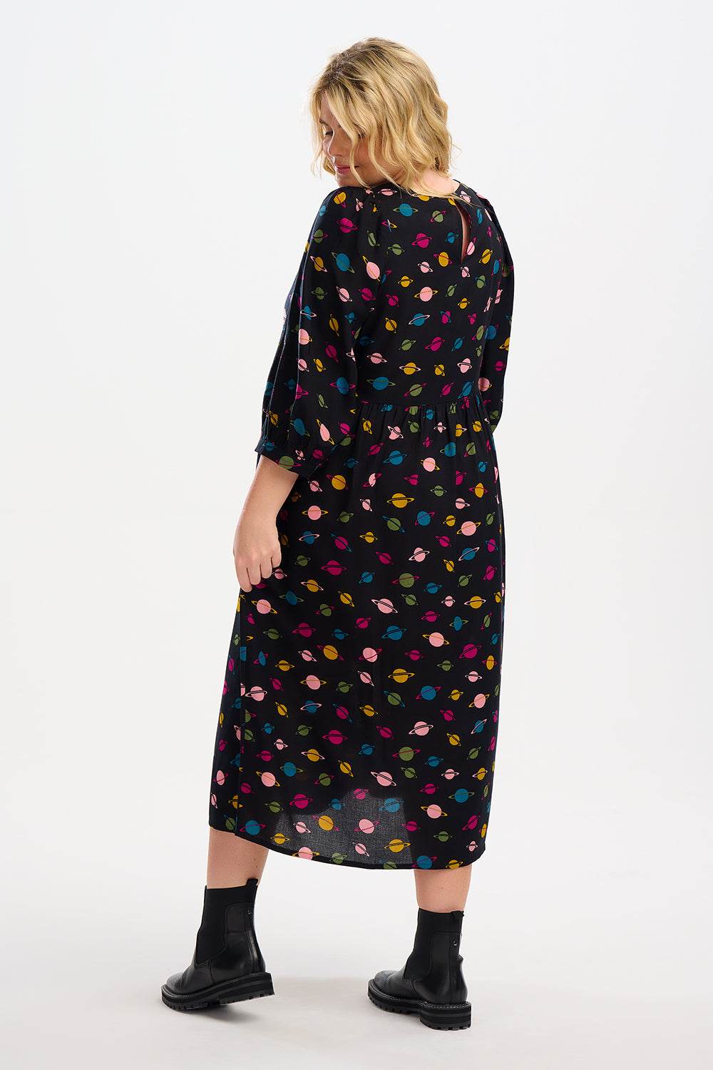A woman standing, wearing a midi smock dress with a colorful planet pattern on a black background, paired with black boots.