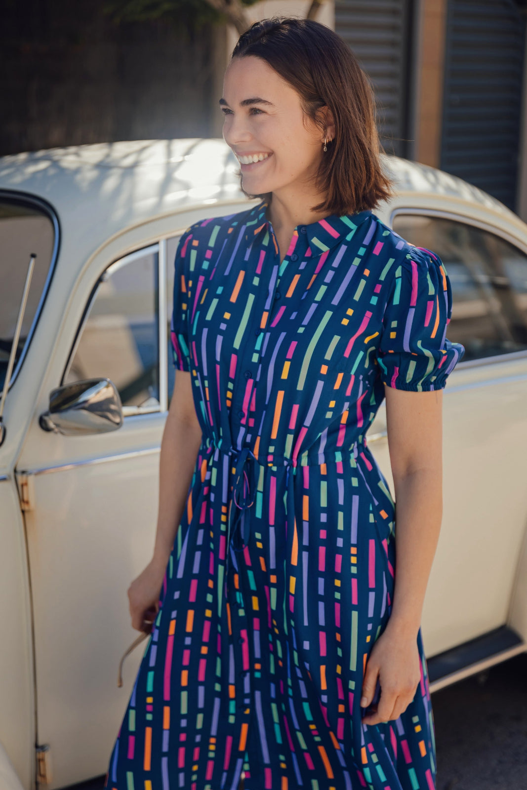 A person smiling and wearing a blue mini shirt dress with a geometric broken lines pattern.