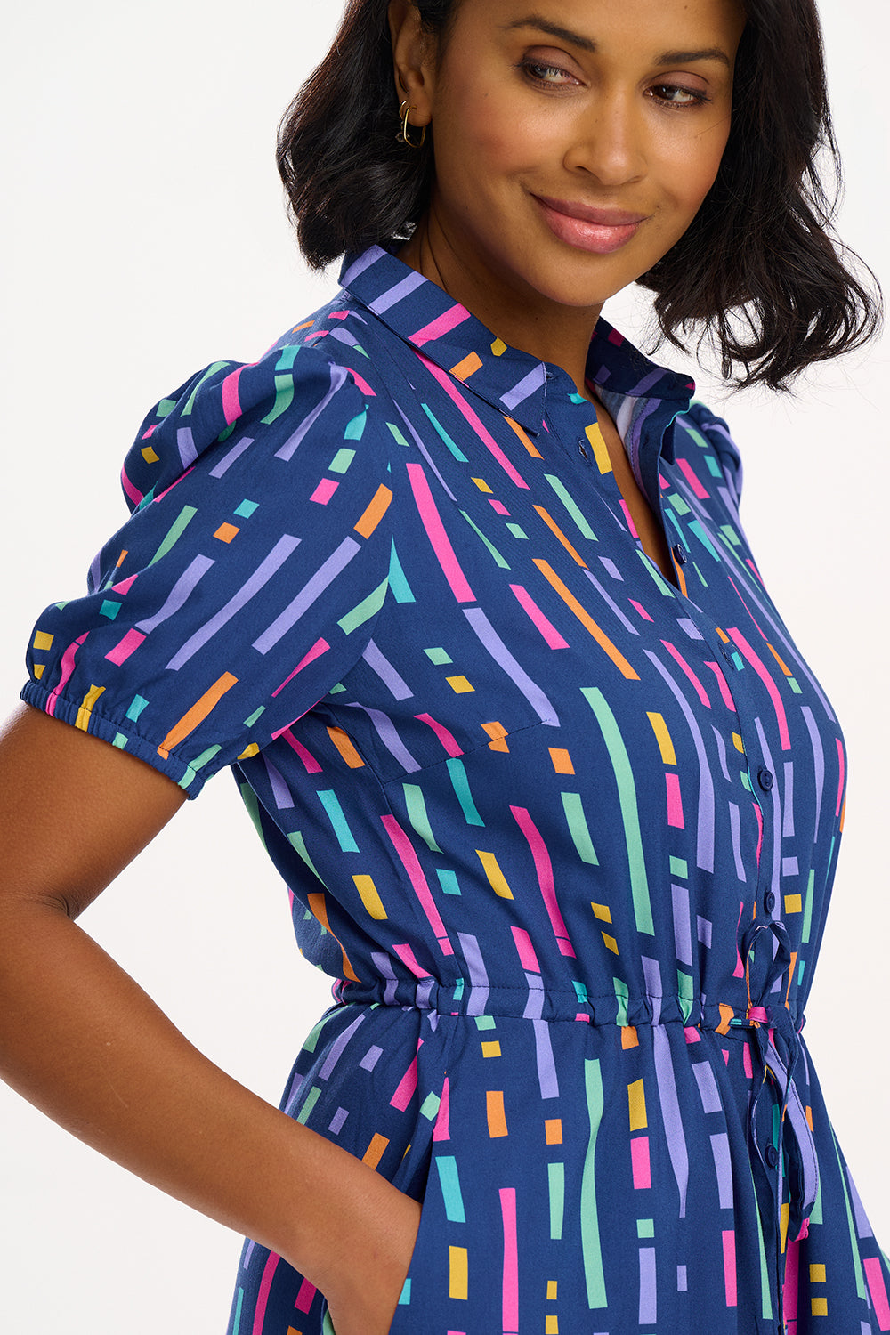 A model wearing a short-sleeved mini shirt dress with a Broken Lines print in blue, pink, purple, and orange on a dark teal blue base. The dress features a button-up front, a soft shirt collar, and a drawstring waist.