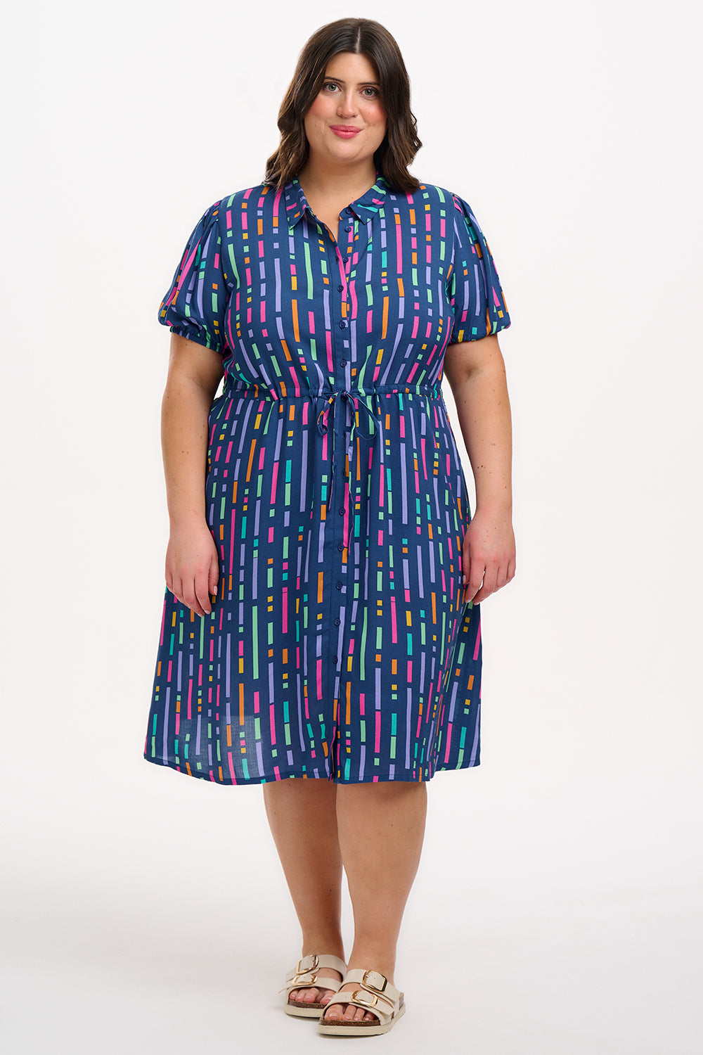 A model wearing a short-sleeved mini shirt dress with a Broken Lines print in blue, pink, purple, and orange on a dark teal blue base. The dress features a button-up front, a soft shirt collar, and a drawstring waist.