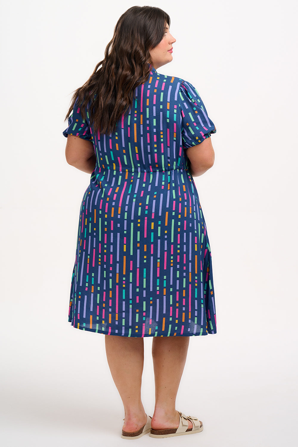 A model wearing a short-sleeved mini shirt dress with a Broken Lines print in blue, pink, purple, and orange on a dark teal blue base. The dress features a button-up front, a soft shirt collar, and a drawstring waist.