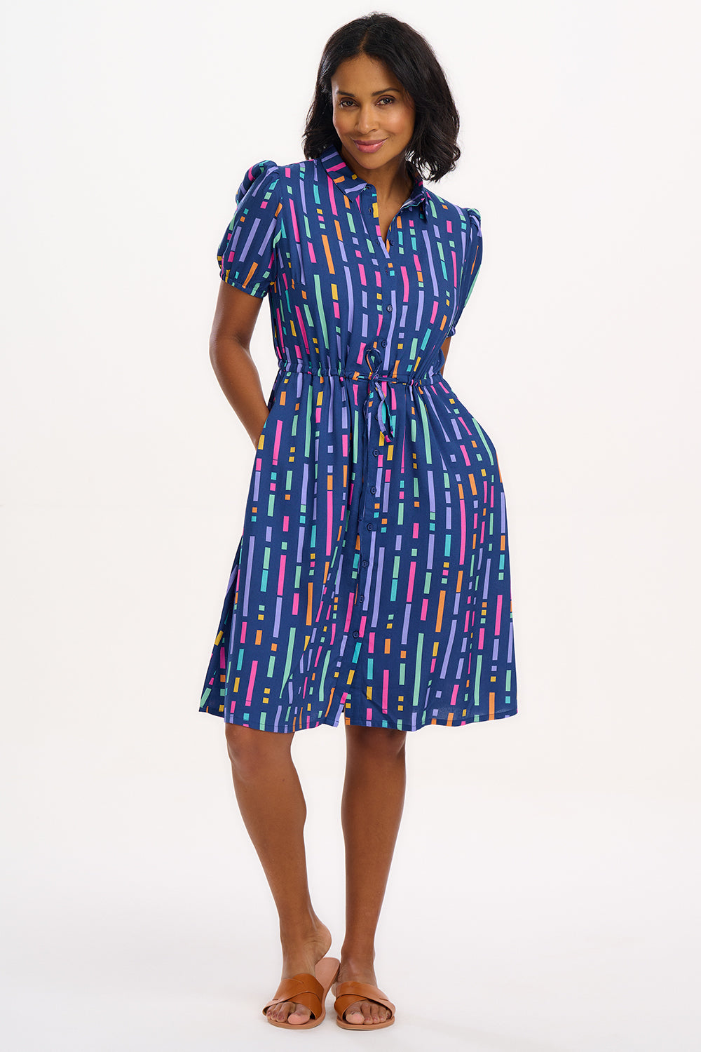 A model wearing a short-sleeved mini shirt dress with a Broken Lines print in blue, pink, purple, and orange on a dark teal blue base. The dress features a button-up front, a soft shirt collar, and a drawstring waist.