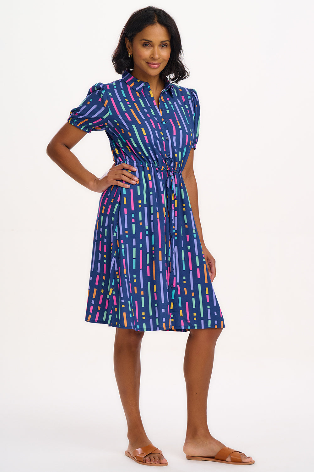 A model wearing a short-sleeved mini shirt dress with a Broken Lines print in blue, pink, purple, and orange on a dark teal blue base. The dress features a button-up front, a soft shirt collar, and a drawstring waist.
