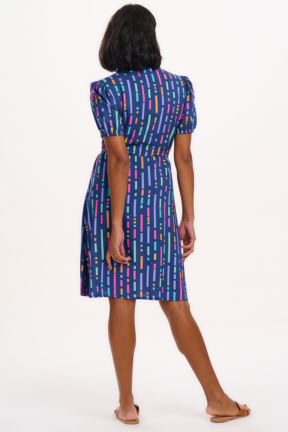 A model wearing a short-sleeved mini shirt dress with a Broken Lines print in blue, pink, purple, and orange on a dark teal blue base. The dress features a button-up front, a soft shirt collar, and a drawstring waist.