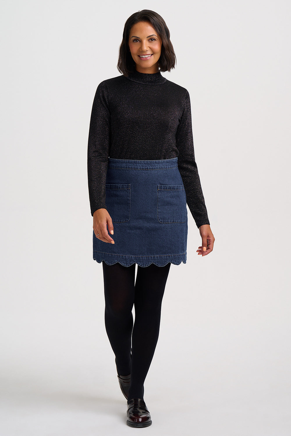 A woman modeling a dark blue denim mini skirt with a scalloped hem, paired with a black long-sleeve knit top, black tights, and black shoes, against a plain light gray studio background.