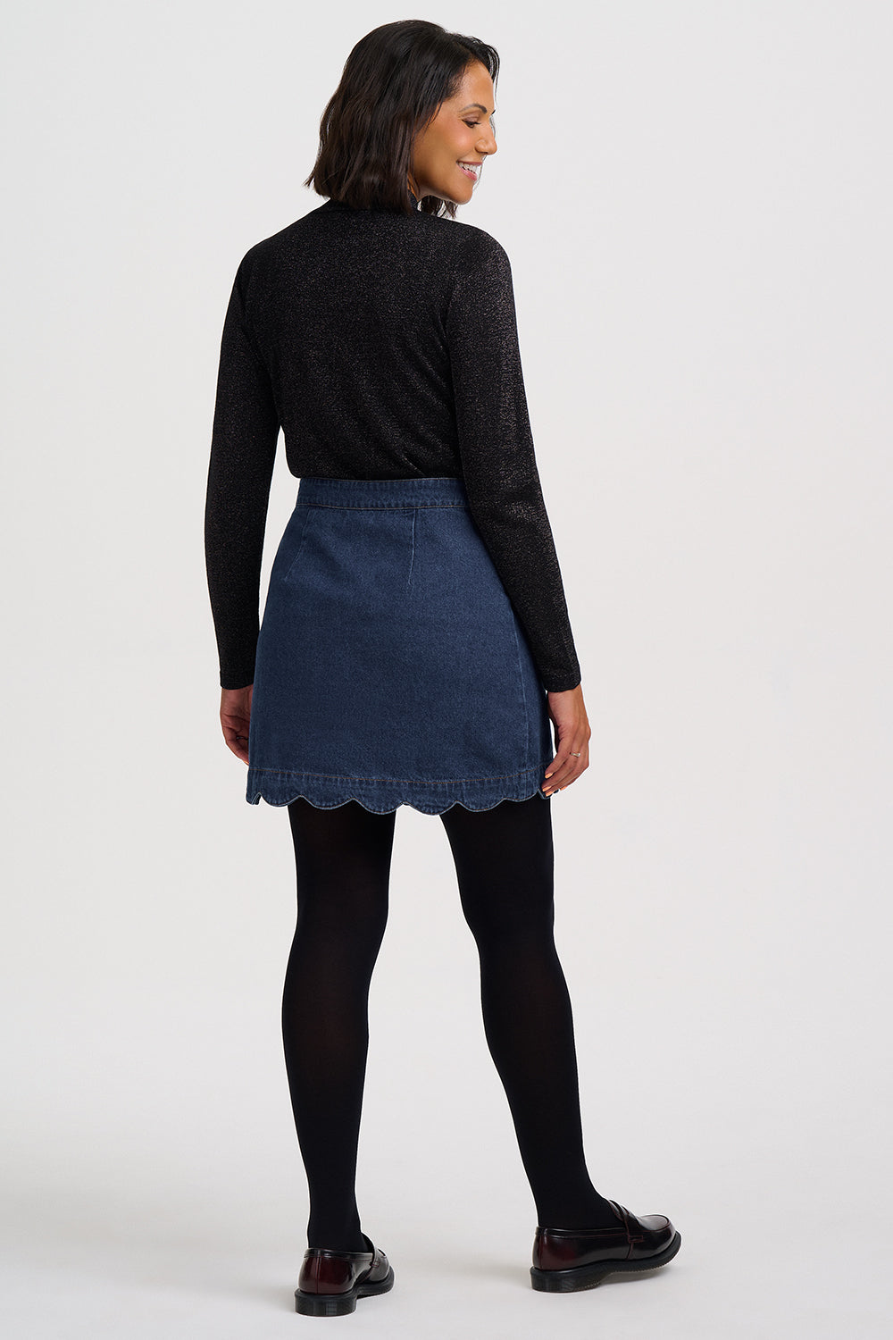 A woman shown from the back wearing a black long-sleeve knit top, a dark blue denim mini skirt with a scalloped hem, black tights, and dark brown leather shoes, against a plain light gray studio background.