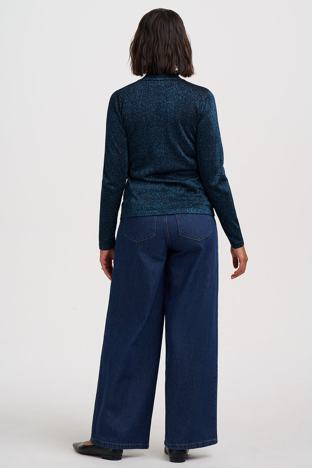 A model shown from the back wearing a dark teal textured knit long-sleeve top and dark blue wide-leg jeans against a plain light gray studio background.