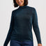 A woman wearing a dark teal long-sleeve mock neck knit sweater and dark blue jeans, standing against a light gray studio background.