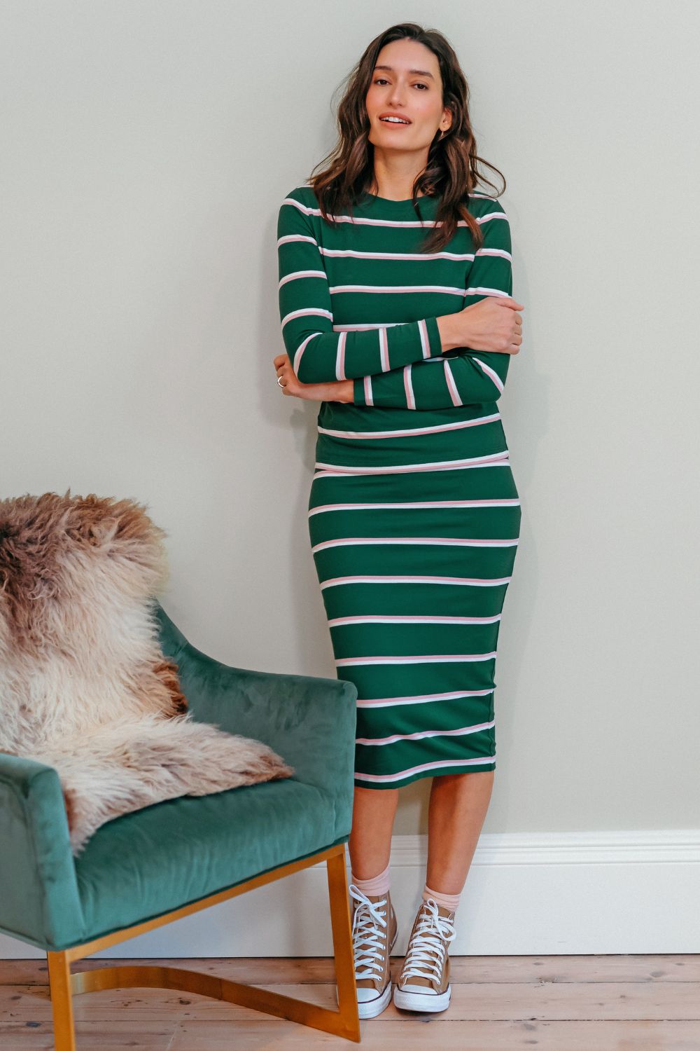 Sugarhill brighton model wearing a green and white striped dress standing in a room with a green chair and wooden floor.