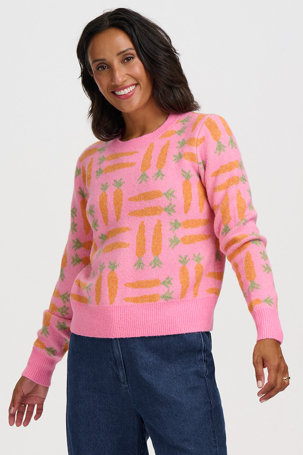 A woman wearing a pink crewneck sweater with an all-over orange and green carrot pattern, paired with dark blue jeans, against a light gray studio background.