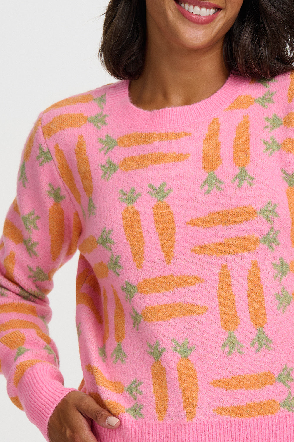 A close-up of a woman wearing a pink knit sweater with an allover carrot pattern in orange and green, against a plain light background.
