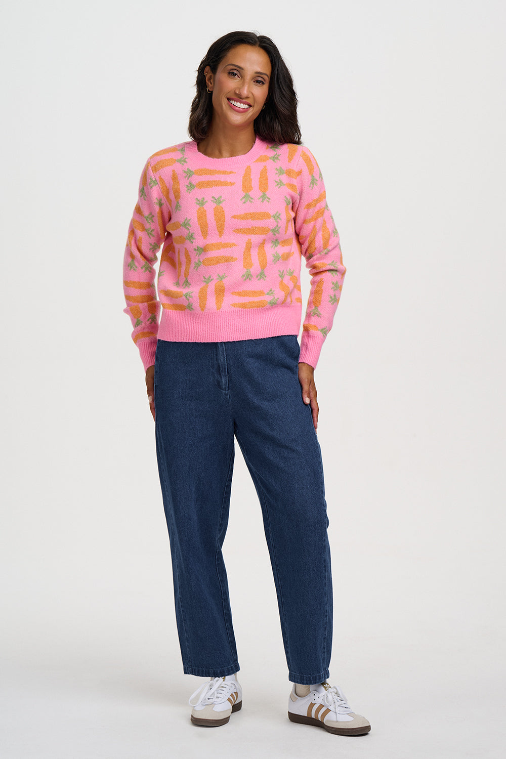 A woman modeling a pink crewneck sweater with an orange and yellow abstract pattern, paired with dark blue jeans and white sneakers, against a light gray studio background.