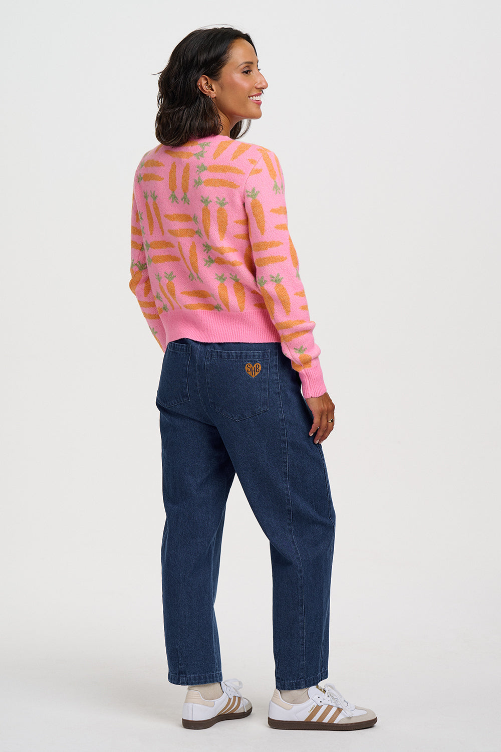 A woman is shown from the back wearing a pink crewneck sweater with a yellow and orange pineapple pattern, dark blue jeans, and white sneakers with gold stripes, against a light gray studio background.