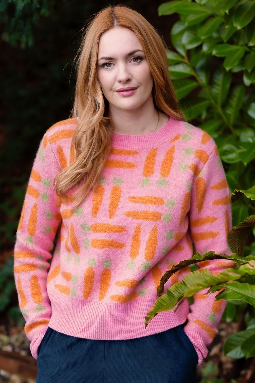 A woman with long red hair wearing a pink knit sweater with an orange and green carrot pattern, standing outdoors in front of green foliage.