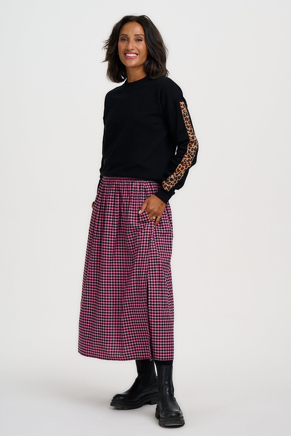 A woman modeling a black long-sleeve top with leopard-print sleeve details and a high-waisted, knee-length burgundy and black gingham skirt, paired with black boots, against a plain light gray studio background.