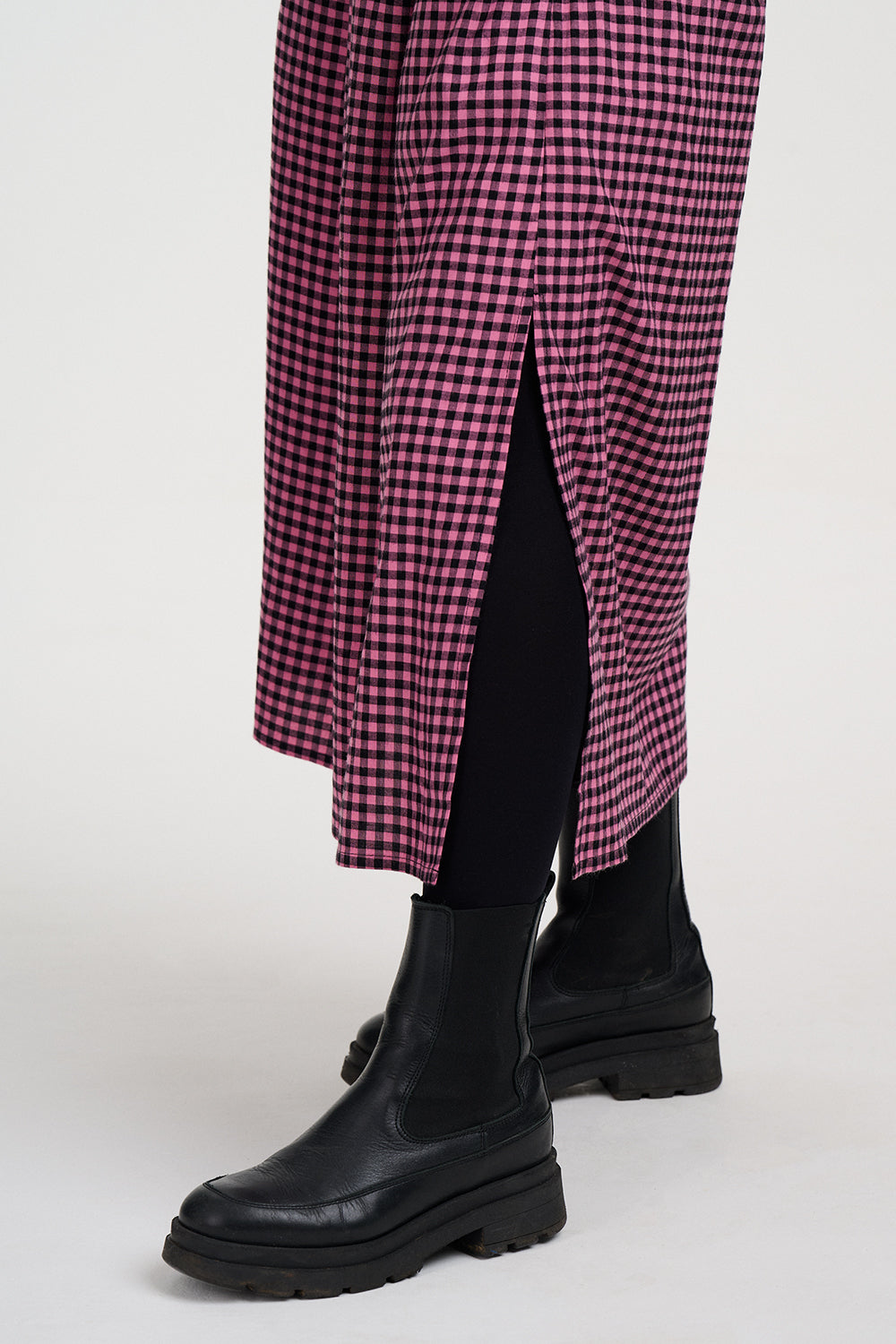 A close-up of a person's legs wearing a pink and black gingham skirt with a side slit, paired with black leggings and chunky black boots, against a light gray studio background.