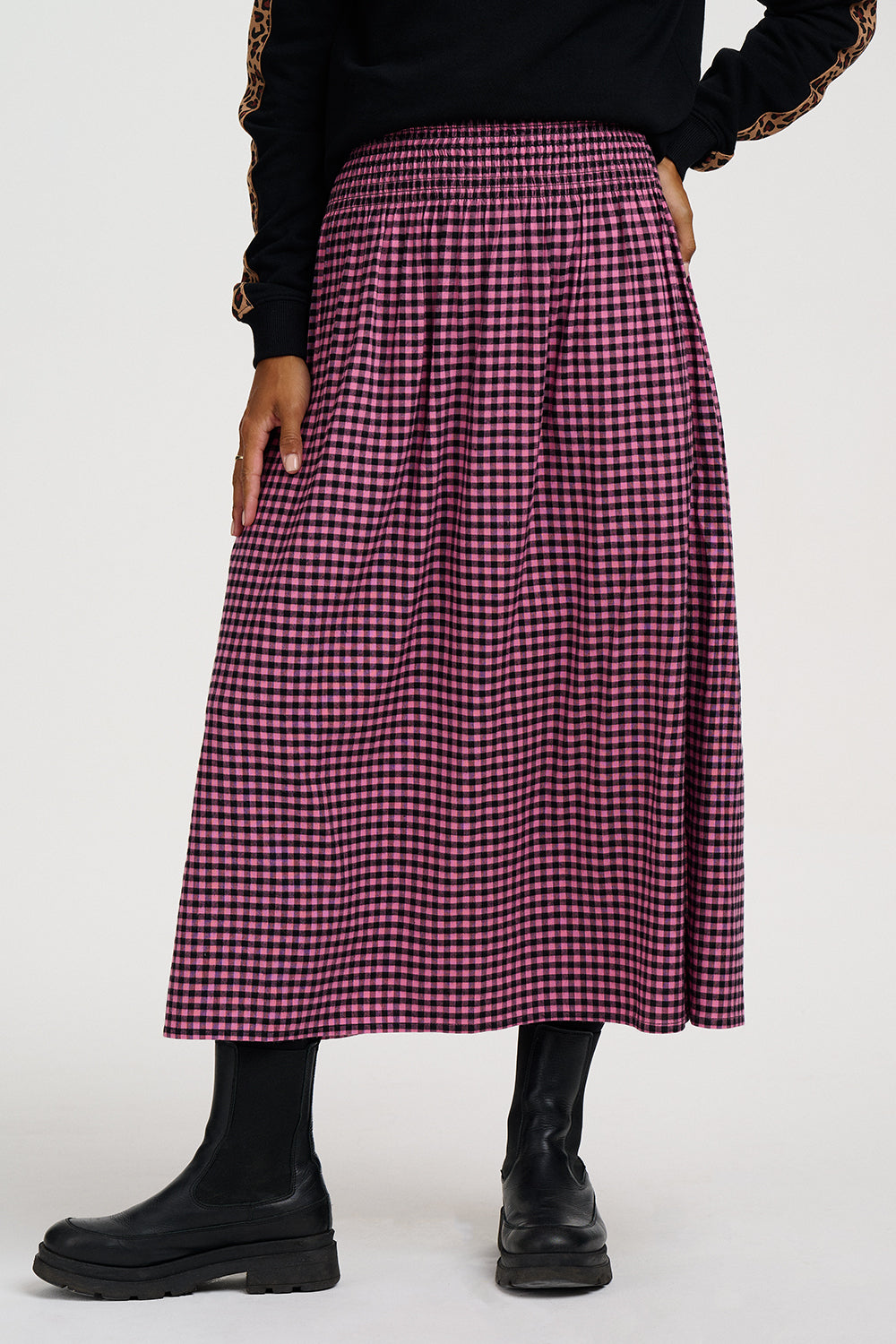 A model wearing a high-waisted, pleated A-line midi skirt in a dark pink and black gingham pattern, styled with a black long-sleeve top and black leather boots against a plain white studio background.