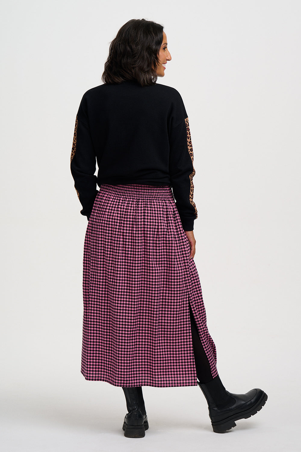 A model is shown from the back wearing a black long-sleeve top with patterned sleeves and a red and black gingham midi skirt with a side slit, paired with black boots, against a plain light gray background.