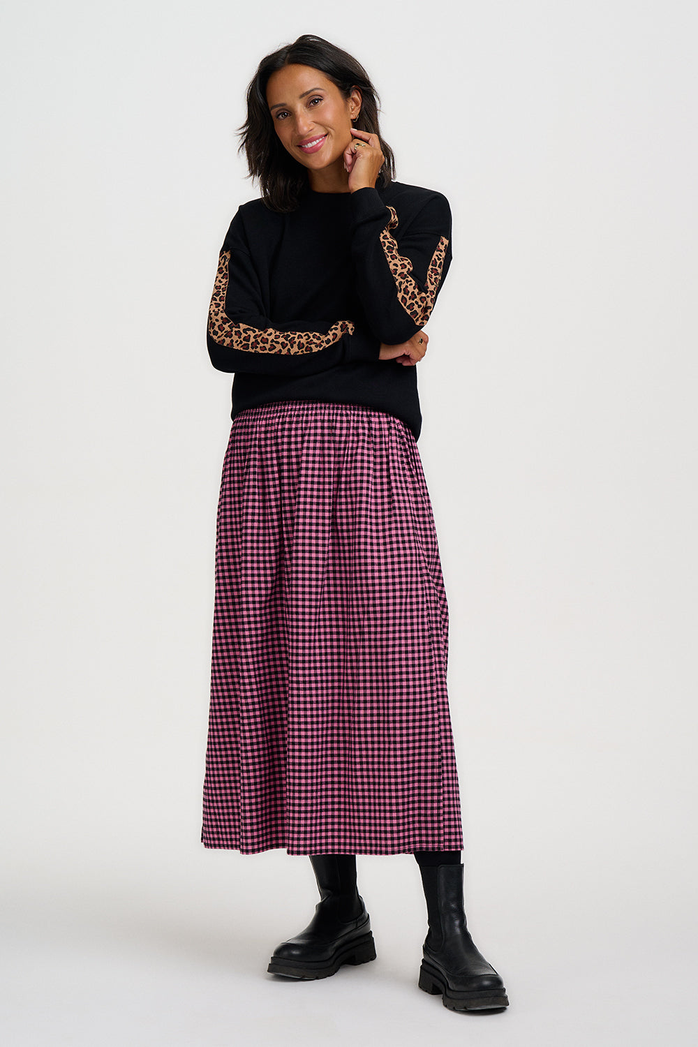 A woman modeling a black long-sleeve top with leopard-print sleeve panels and a high-waisted, red-and-white gingham pleated midi skirt, paired with black boots, against a light gray studio background.
