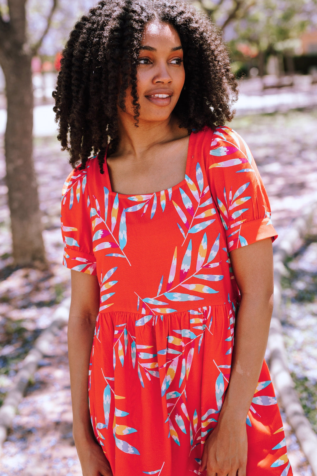 A model wearing a red smock dress with a rainbow fronds print, featuring a square neckline, short sleeves, and a short length.