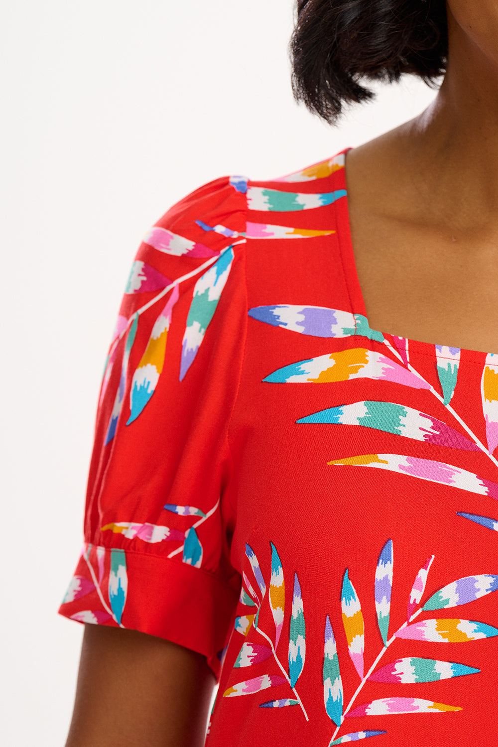 A woman wearing a red smock dress with a Rainbow Fronds print consisting of colorful tropical leaves.