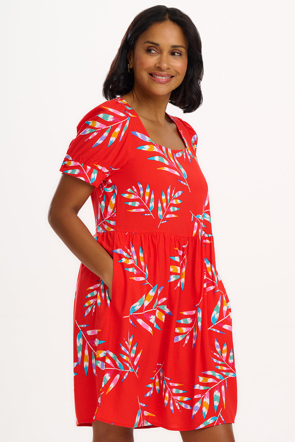A woman wearing a red smock dress with a Rainbow Fronds print consisting of colorful tropical leaves.