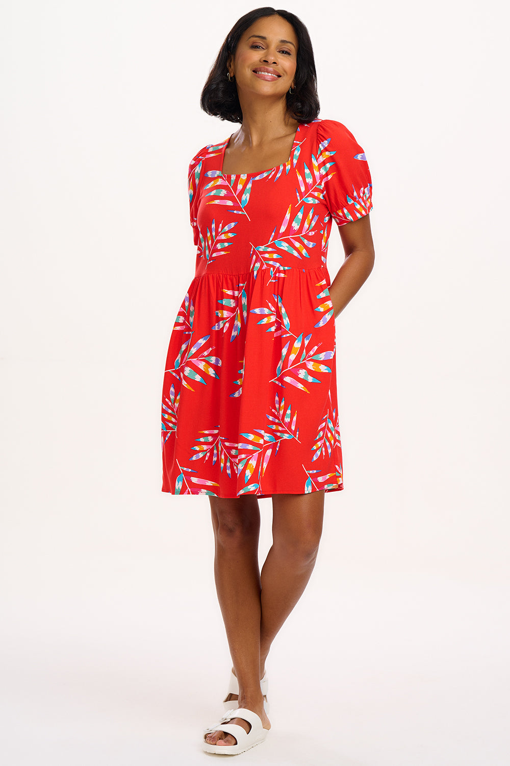 A woman wearing a red smock dress with a Rainbow Fronds print consisting of colorful tropical leaves.