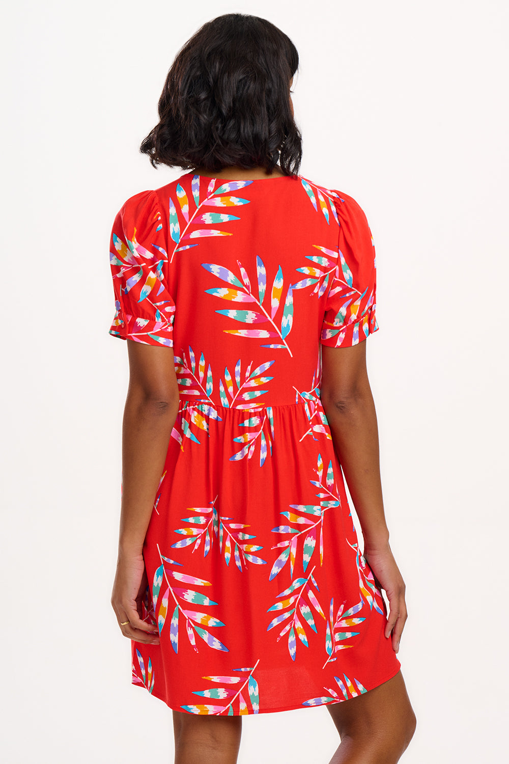 A woman wearing a red smock dress with a Rainbow Fronds print consisting of colorful tropical leaves.