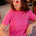 Woman wearing a pink t-shirt with a sun design and heart-shaped sunglasses indoors.