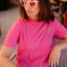 Woman wearing a pink t-shirt with a sun design and heart-shaped sunglasses indoors.
