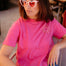 Woman wearing a pink t-shirt with a sun design and heart-shaped sunglasses indoors.