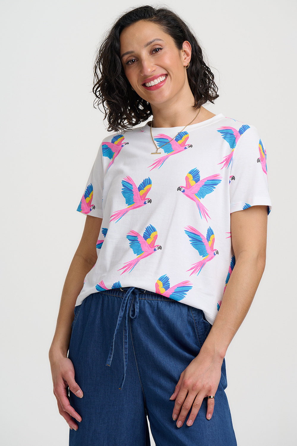 Woman wearing a white t-shirt with colorful parrot patterns and blue pants on a plain background