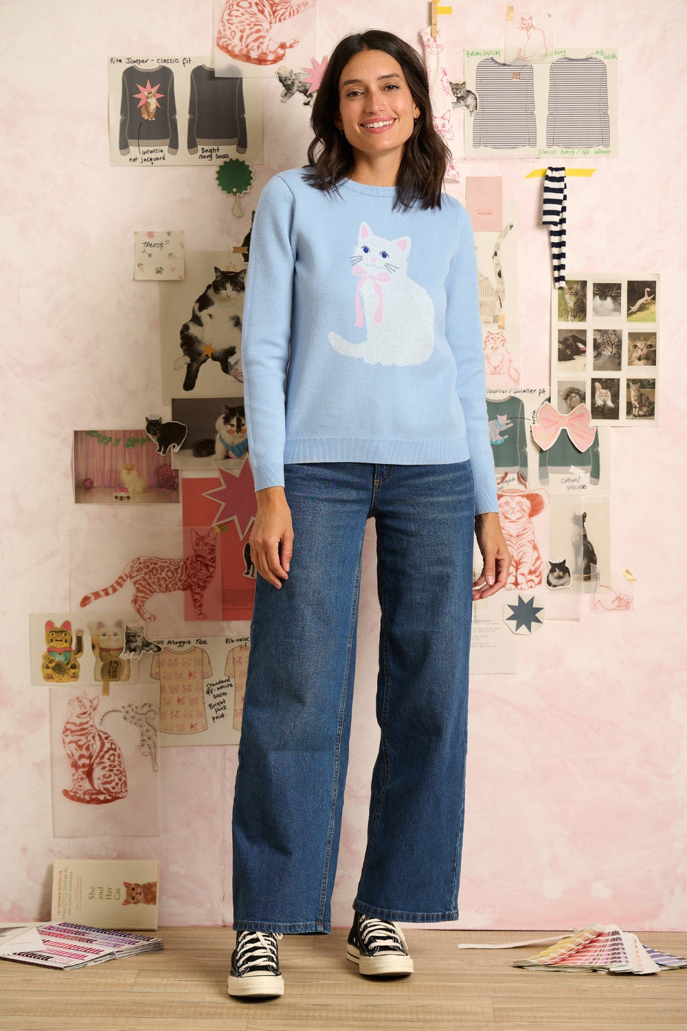 Woman wearing a blue sweater with a cat design in a room with a wall of cat-themed decorations.
